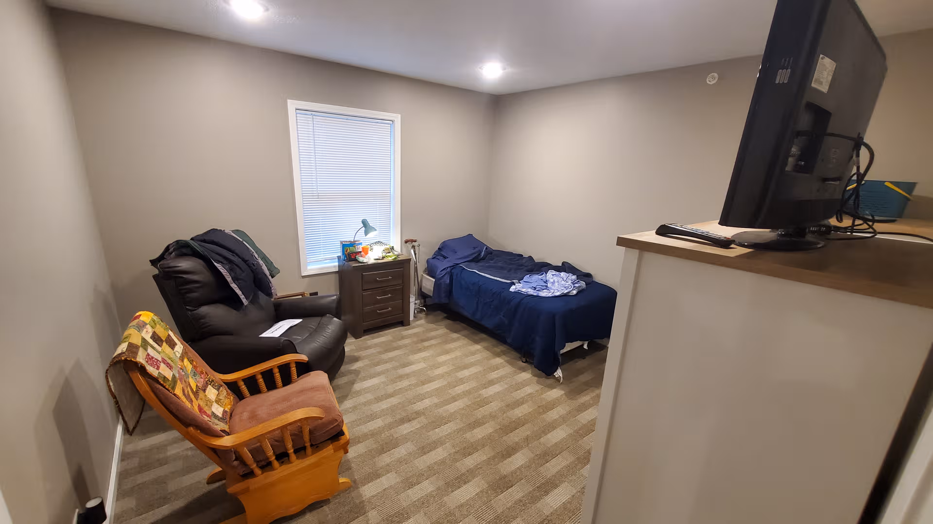 Small furnished bedroom with a bed, recliner, chair, nightstand, and TV