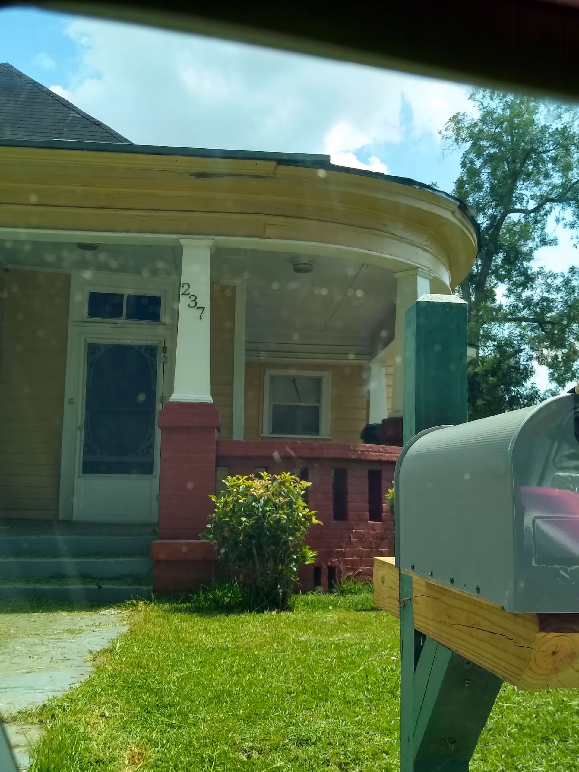 Yellow wooden house front porch with a red brick railing, a mailbox in the foreground, and the house number 237 on a column.
