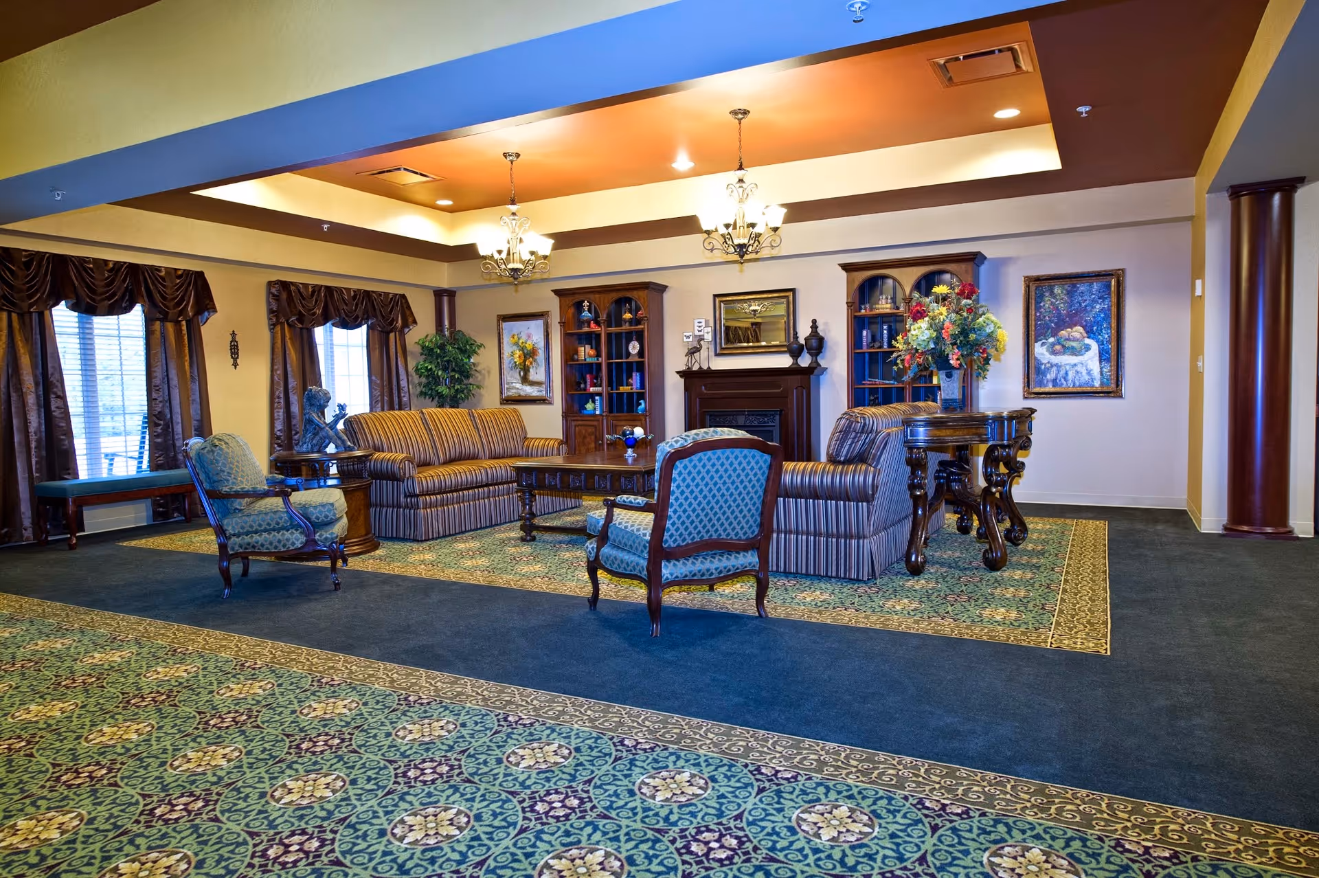 A spacious and elegantly decorated living room area with patterned carpet, two striped sofas, two upholstered armchairs, a wooden coffee table, and a round side table with a large floral arrangement. The room features two wooden bookcases, framed artwork on the walls, a fireplace, and large windows with dark curtains. The ceiling has recessed lighting and two chandeliers.