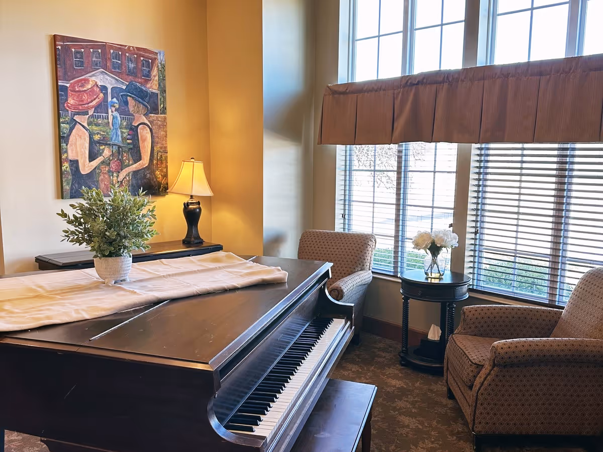 A cozy interior room featuring a grand piano with a white runner and a potted plant on top. Two patterned armchairs are positioned near a window with blinds and a valance. A small round table between the chairs holds a vase with white flowers. A lamp and a colorful painting of two women wearing hats hang on the wall behind the piano.