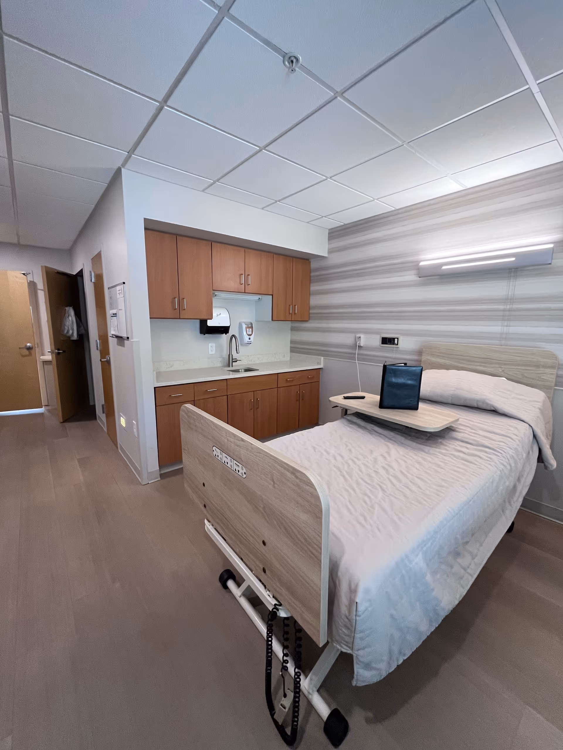 A hospital-style patient room with a bed, wooden cabinets, and a small kitchenette.