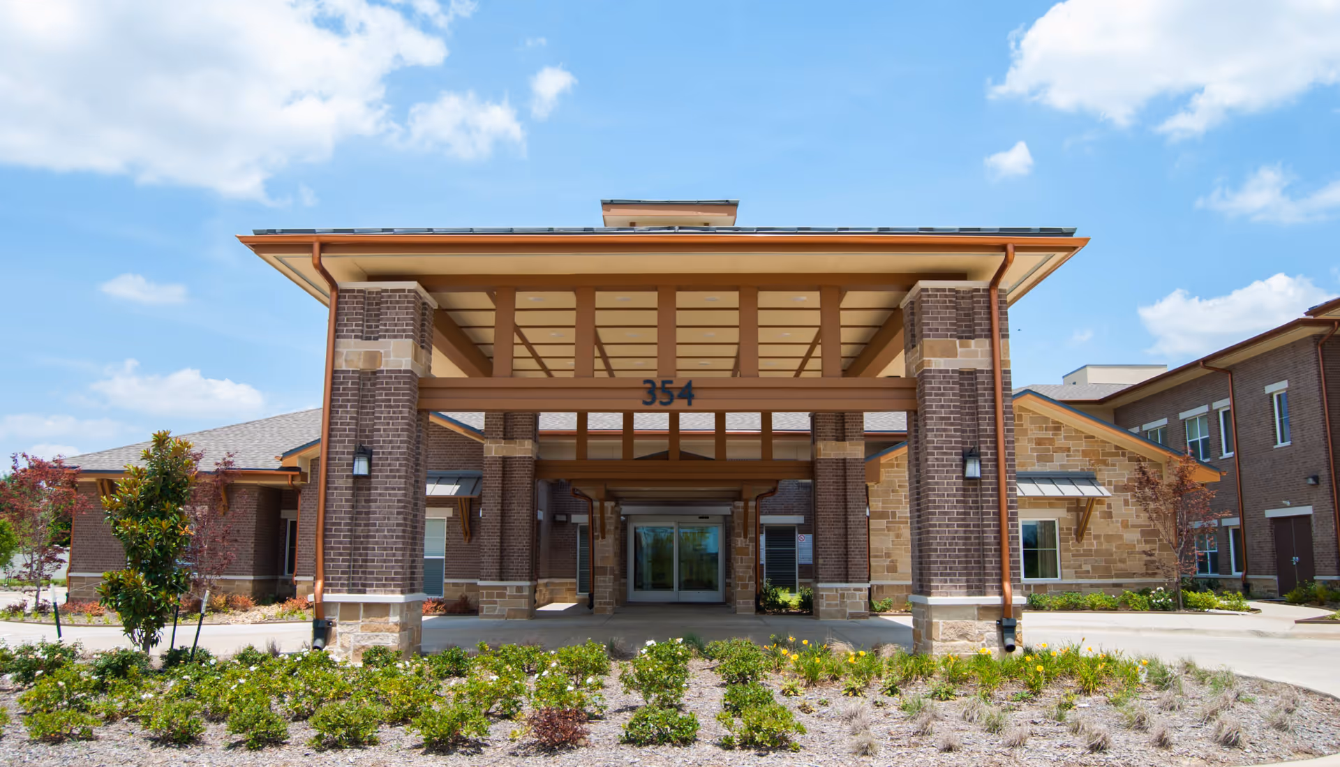 Front exterior view of a senior living facility building with a covered entrance supported by brick columns, landscaped greenery in front, and a clear blue sky with some clouds.