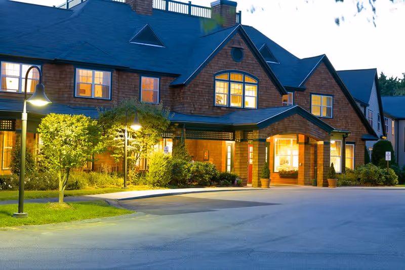 Front entrance of a lit senior living facility with gabled roof, landscaped beds, and street lamps at dusk.