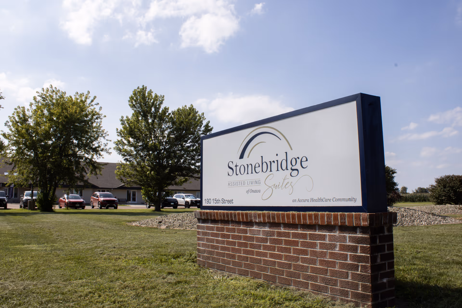 Stonebridge Suites of Onawa exterior with a large brick sign and parked cars.