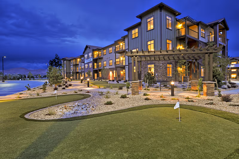 Exterior front of a senior living building at dusk with lit windows, a pergola and a small putting green in the foreground.