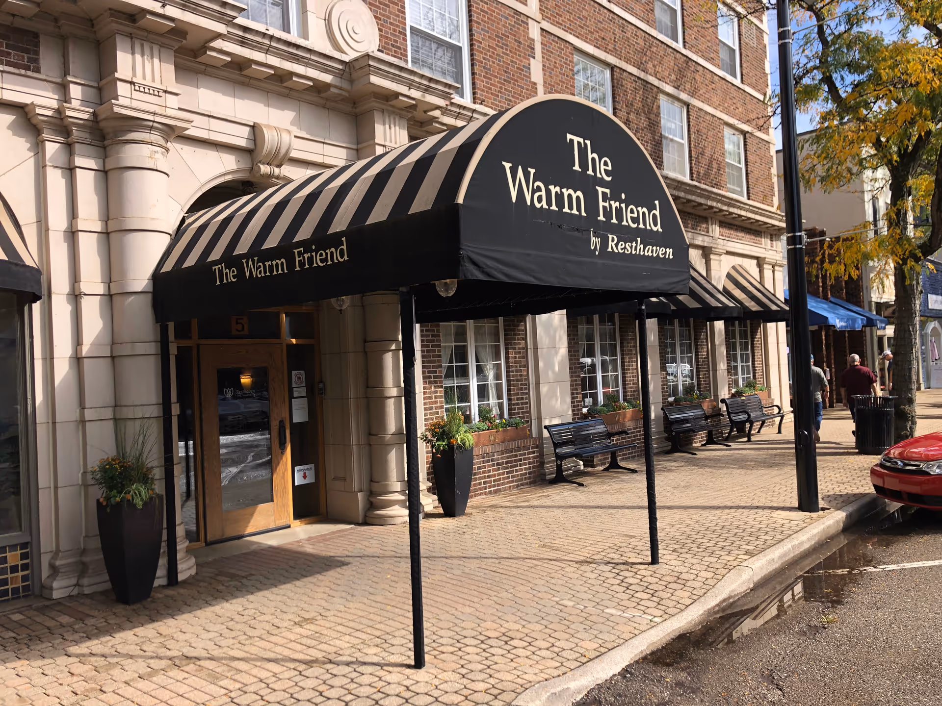Brick building storefront with striped awning reading The Warm Friend, benches and planters along sidewalk.