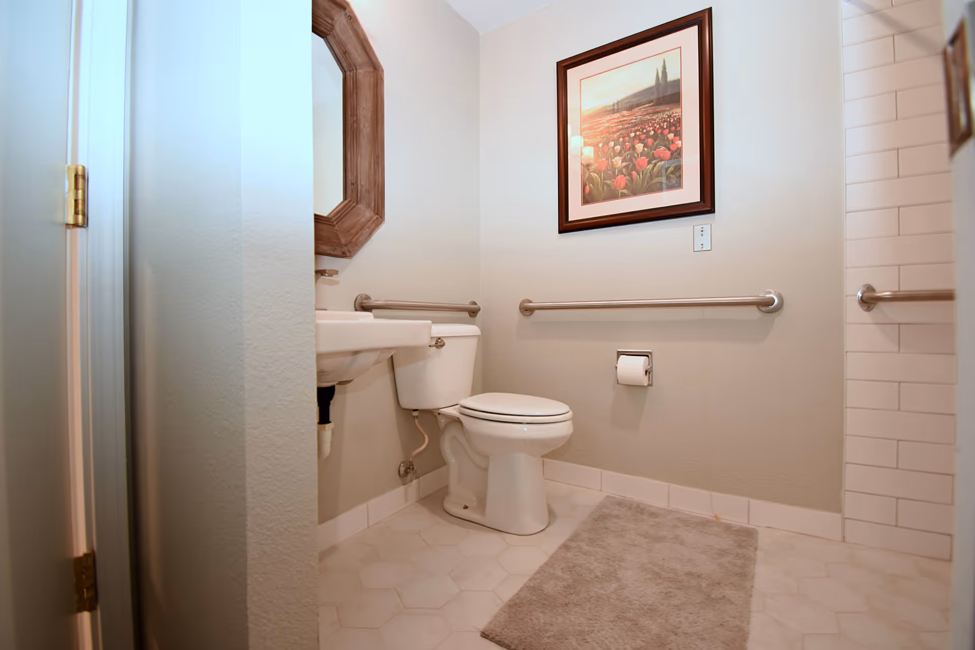A clean and accessible bathroom with a white toilet, a small wall-mounted sink, grab bars on the walls, a beige bath mat on the tiled floor, and a framed picture of a field of flowers hanging above the toilet.