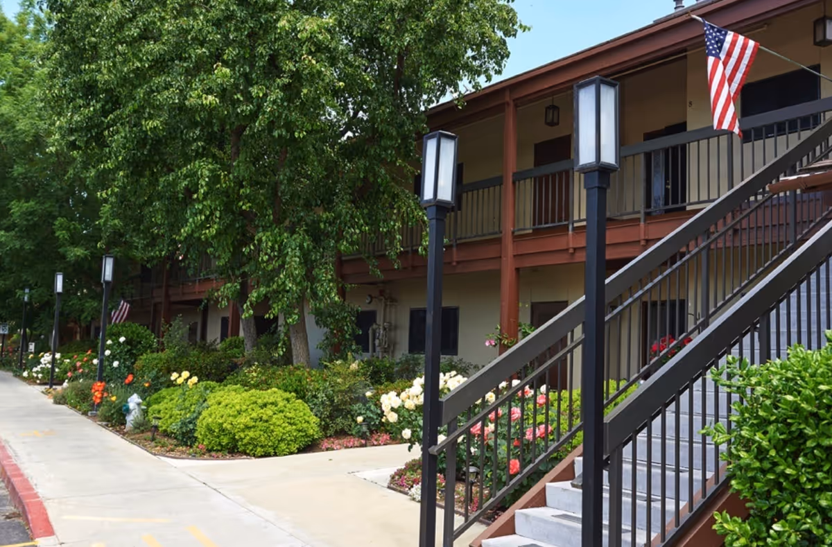 Exterior of a two-story residential building with landscaped gardens, a walkway, stairway with railings, and an American flag.