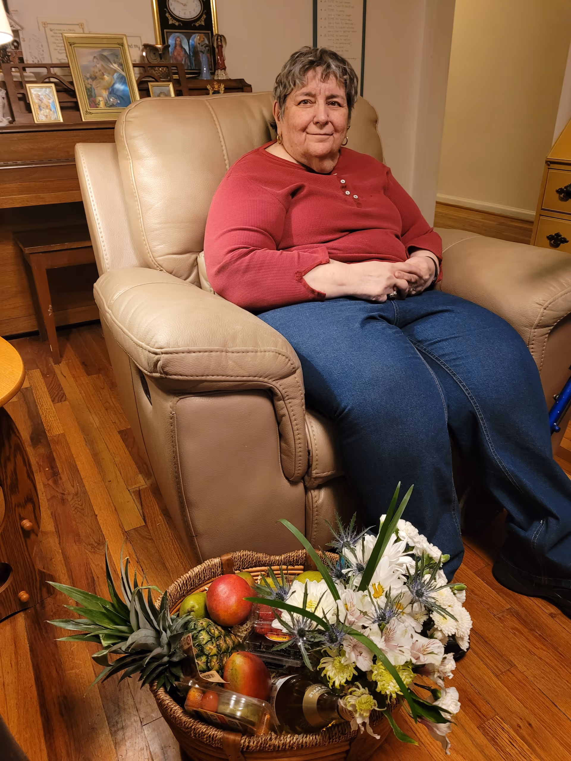 An elderly woman wearing a red long-sleeve shirt and blue jeans is sitting comfortably in a beige leather armchair in a cozy room with wooden floors. In front of her is a wicker basket filled with fresh fruits, flowers, and a bottle. Behind her, there is a piano with framed pictures and a clock on top.
