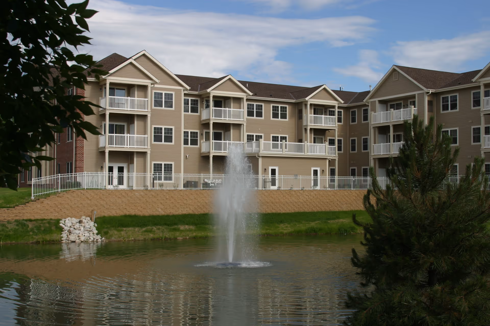 Exterior view of a multi-story residential building with beige siding and white balconies, overlooking a pond with a water fountain in the center. Trees and greenery surround the pond under a partly cloudy sky.