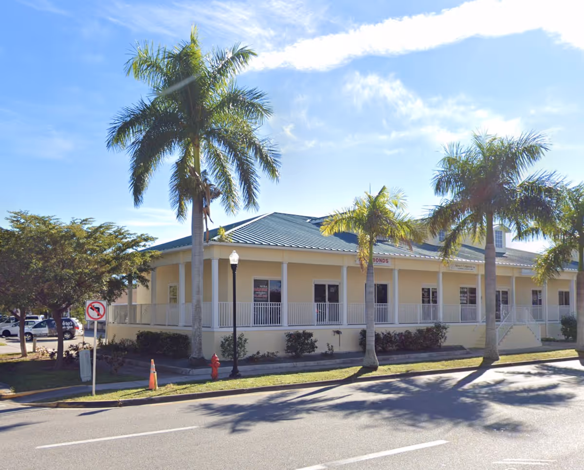 Exterior view of a single-story beige senior living building with a covered porch and palm trees.