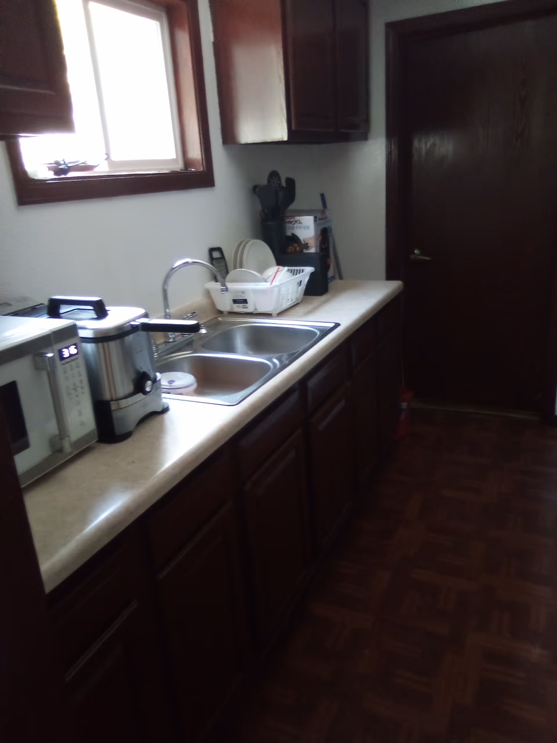 Compact kitchen with sink, dish rack, microwave, coffee maker, dark cabinets, and a window.