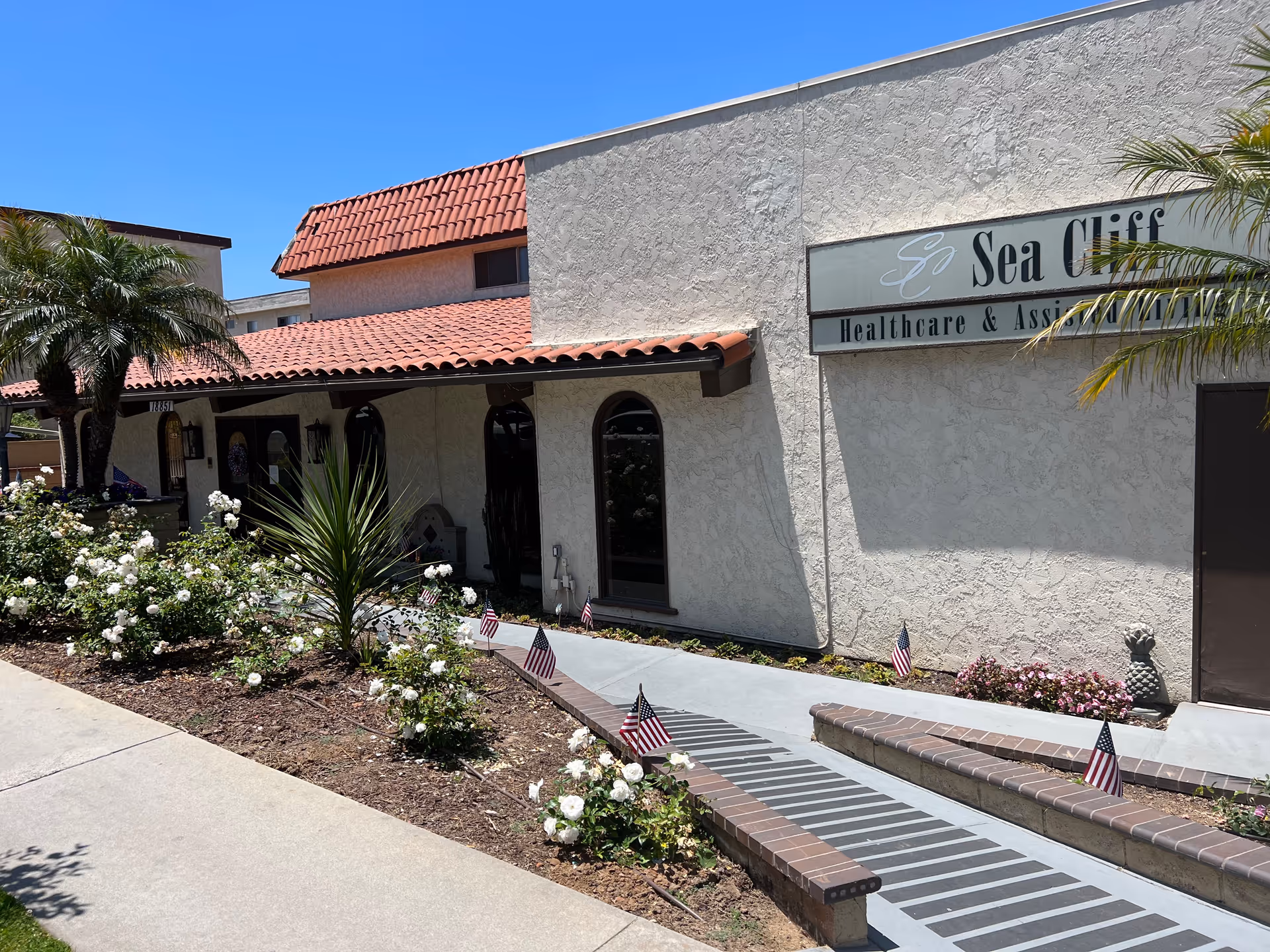 Exterior view of Sea Cliff Assisted Living building with a walkway, plants, flags, and signage.