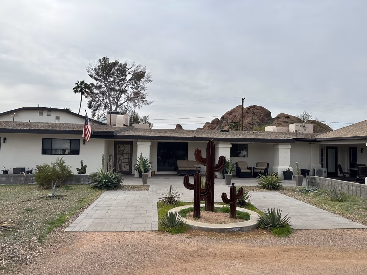 Front exterior of a single-story ranch-style senior living building with a paved walkway, patio seating, an American flag, and metal cactus sculptures in a central planter.