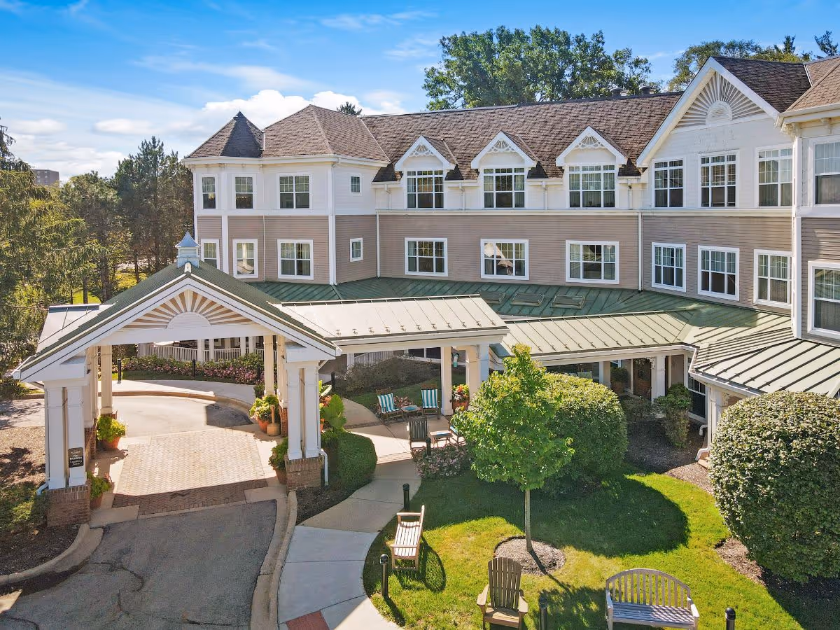Front entrance and covered porte-cochère of a multi-story senior living building with a landscaped courtyard, benches, and seating.