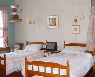 A small bedroom with two single wooden beds, each made with pink and white plaid bedding. Between the beds is a small table holding a small black television and some books. The walls are light-colored with a subtle pattern, decorated with two framed pictures. There are wall-mounted lamps above each bed and patterned curtains on the windows.