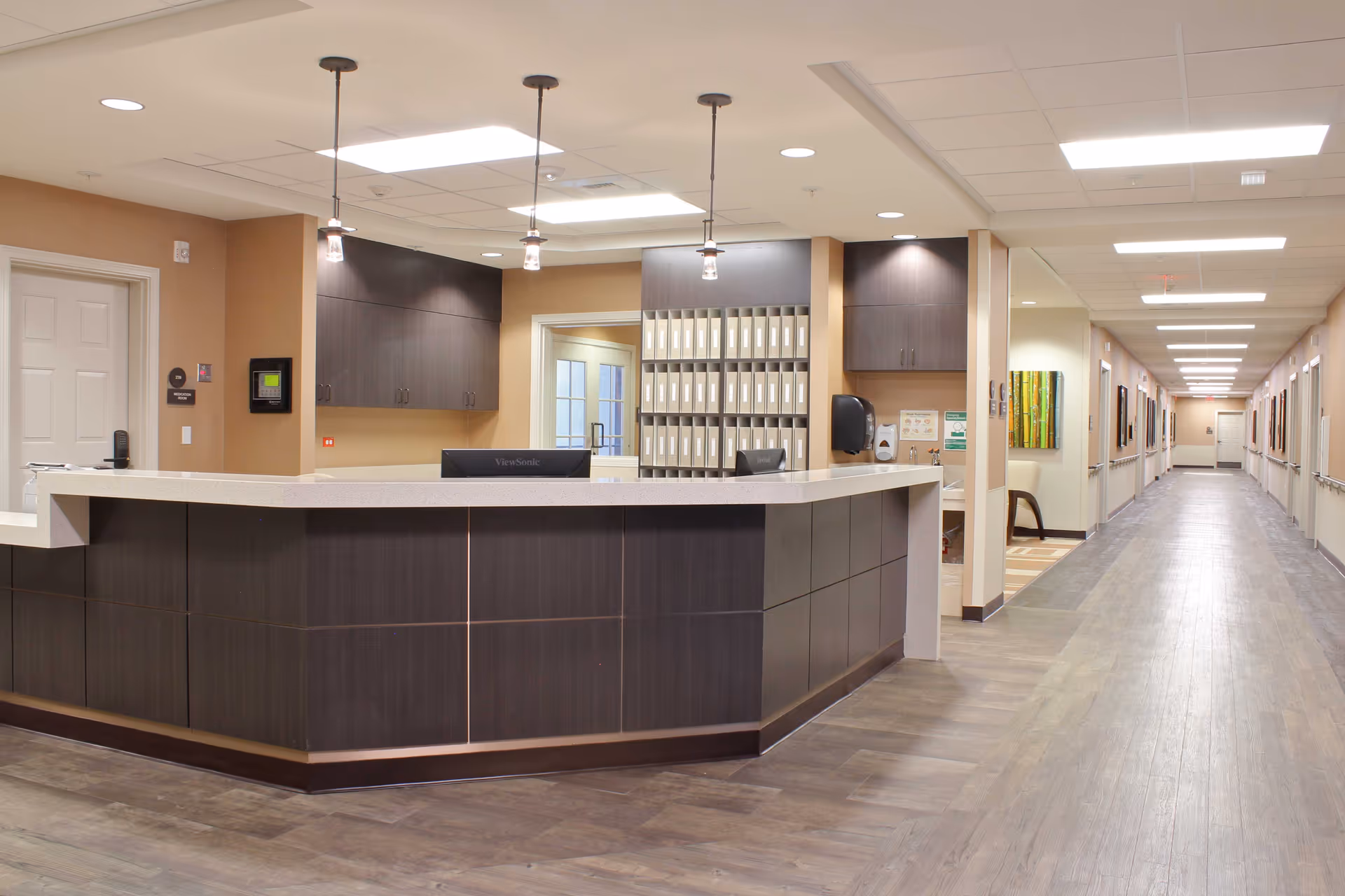 A modern reception desk area inside The Ellison John Transitional Care Center with dark wood cabinetry, a light-colored countertop, and three pendant lights hanging from the ceiling. Behind the desk are shelves with organized files and a hallway extending to the right with handrails and framed artwork on the walls.