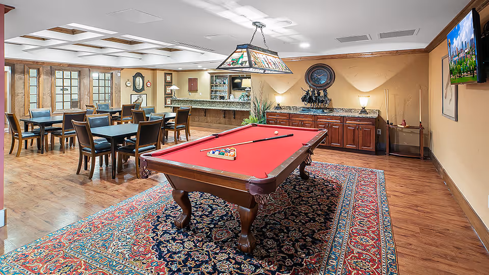 A spacious recreational room featuring a red felt pool table on a patterned rug in the center, surrounded by several tables and chairs. The room has wooden flooring, a decorative ceiling light above the pool table, a wall-mounted TV, and a sideboard with lamps and decor against a beige wall.