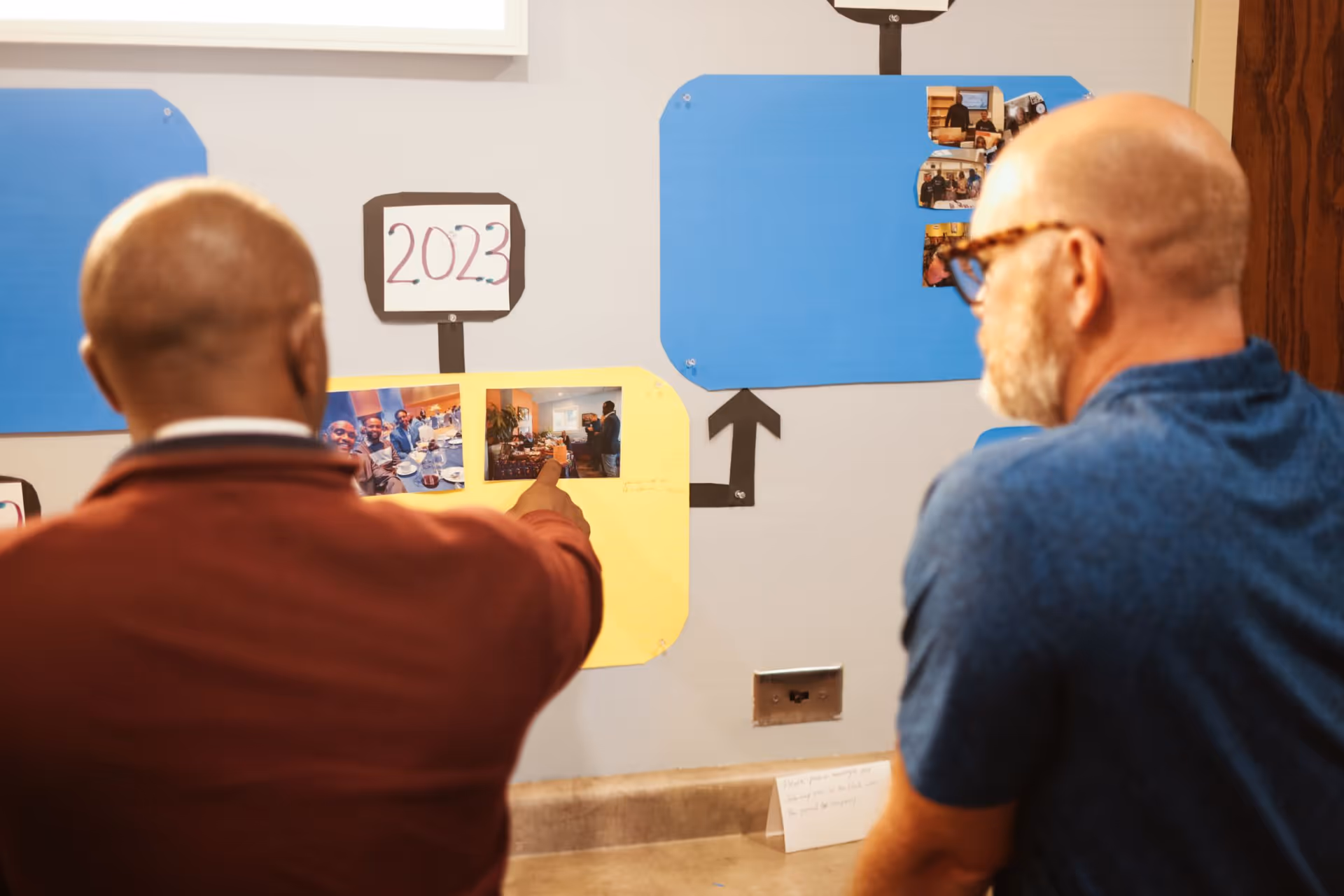 Two men stand with their backs to the camera, looking at a wall display of photos.