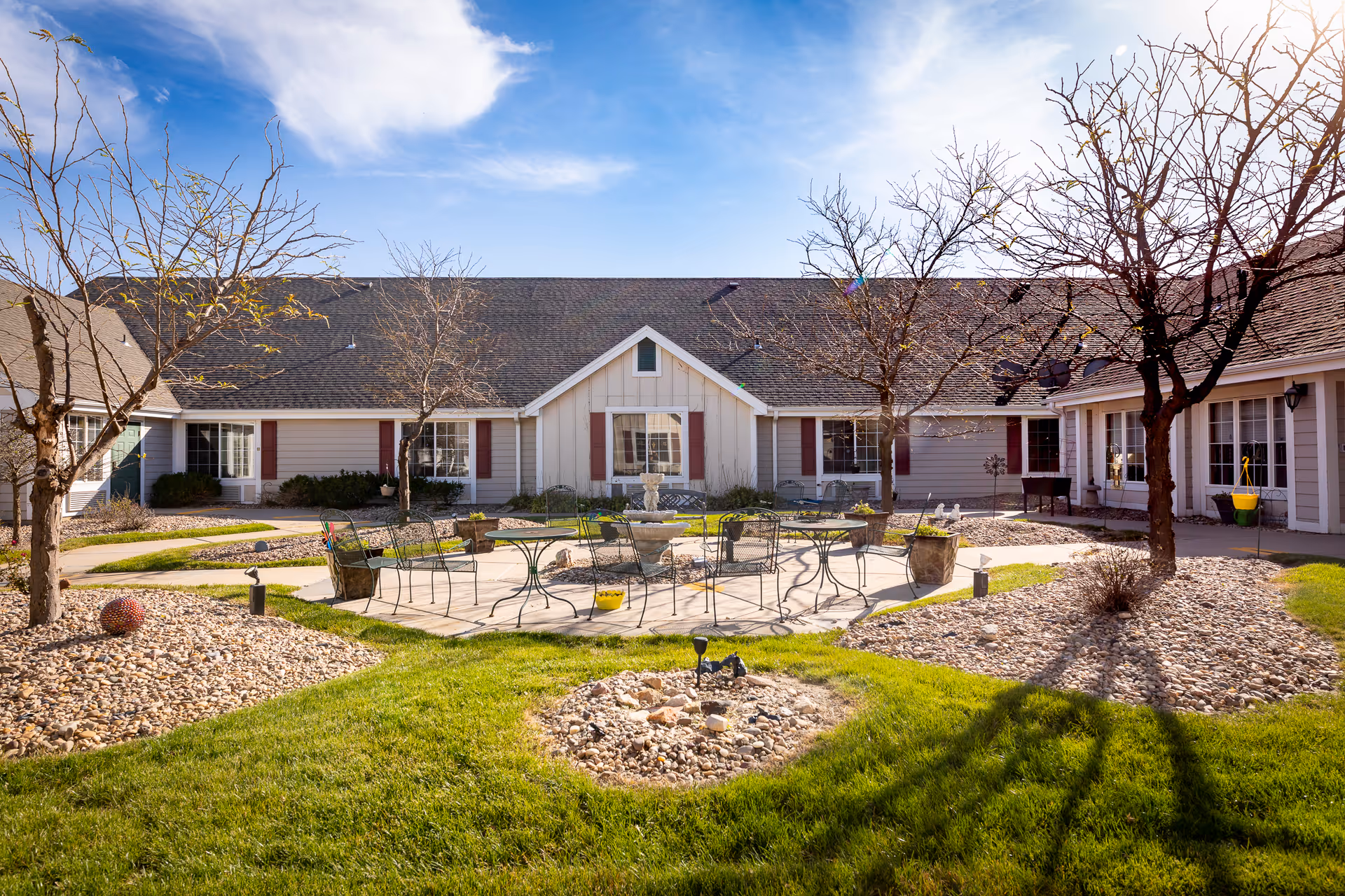 Outdoor courtyard area of Homestead Assisted Living of Hastings featuring a circular paved patio with metal tables and chairs, surrounded by landscaped grass, trees without leaves, and rock beds. The building with beige siding and red shutters encloses the courtyard under a blue sky with some clouds.