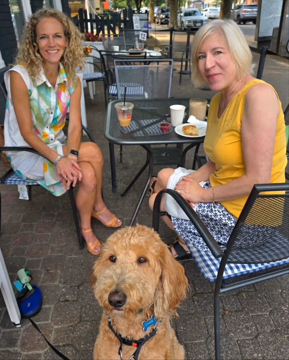 Two women sit at outdoor cafe tables with a golden doodle in the foreground.