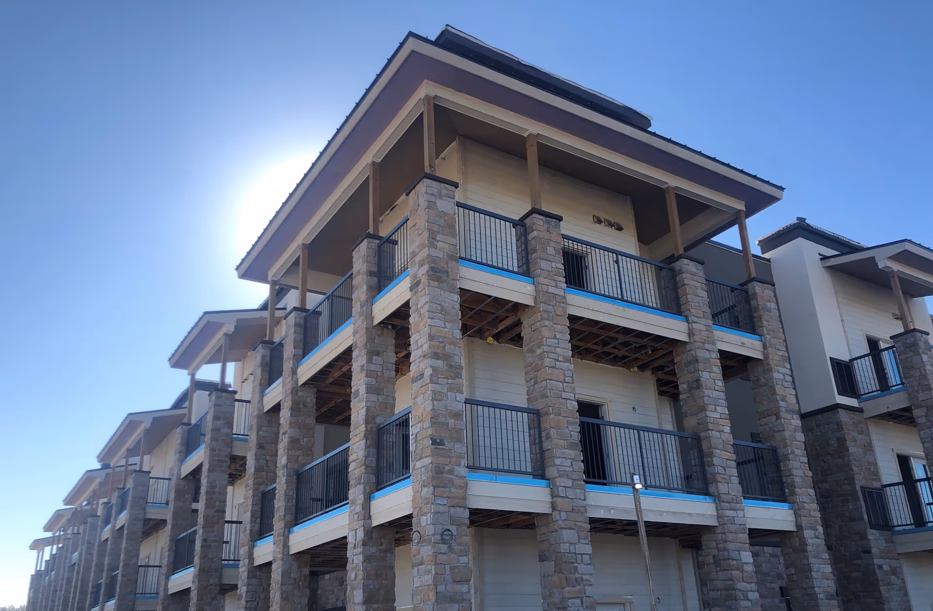 Exterior view of a multi-story building with stone pillars and balconies, under a clear blue sky with the sun shining behind the building.