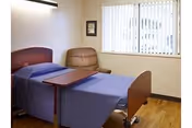 A small bedroom in a senior facility with a bed, chair, and window blinds.