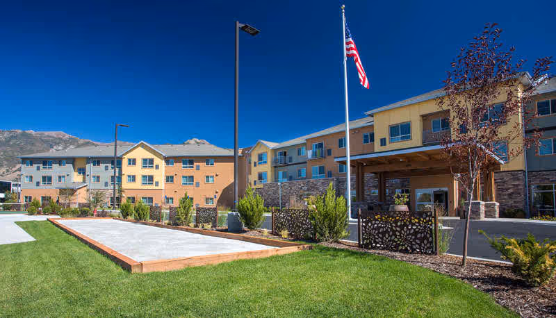 Front exterior of a multi-story senior living building with landscaped lawn, flagpole, and outdoor activity courts against a mountain backdrop.