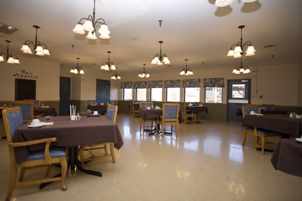 A spacious dining room in a senior living facility with brown tablecloths and chandeliers.