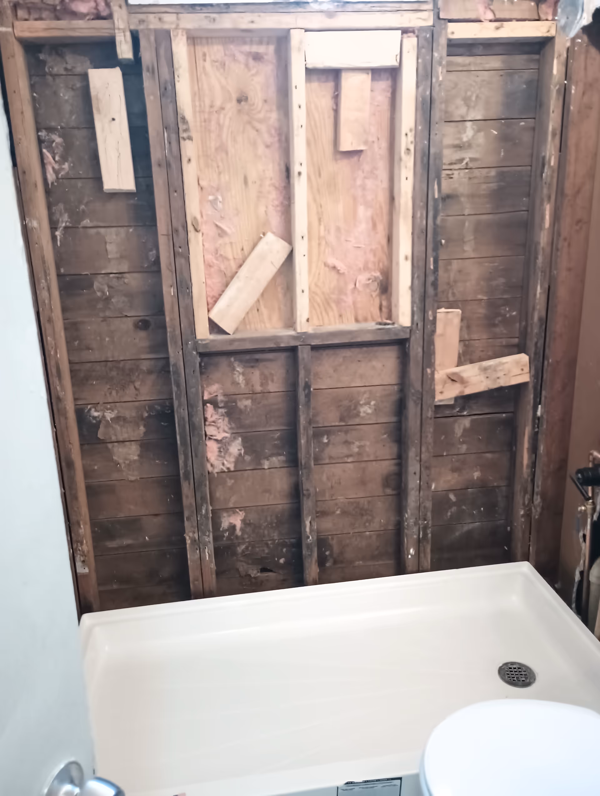 Exposed studs and pink insulation behind a white tub in a bathroom under renovation.