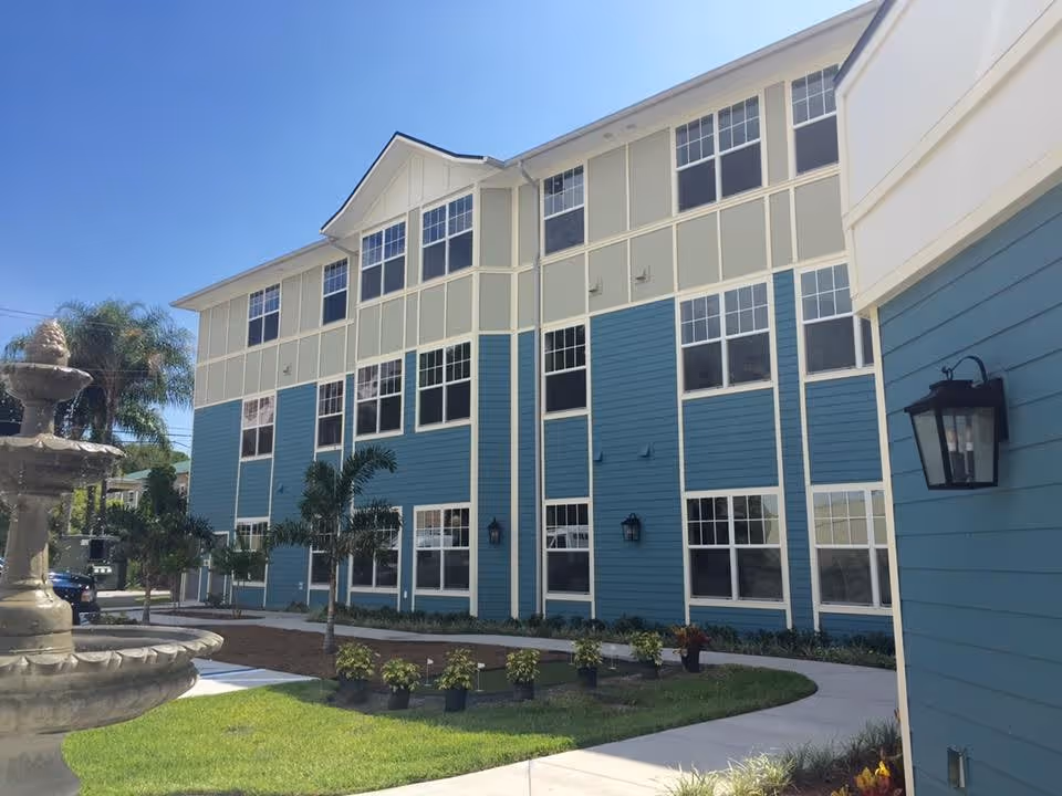 Exterior view of a three-story senior living facility building with blue and beige siding, multiple windows, a paved walkway, small palm trees, and a decorative water fountain in the foreground under a clear blue sky.