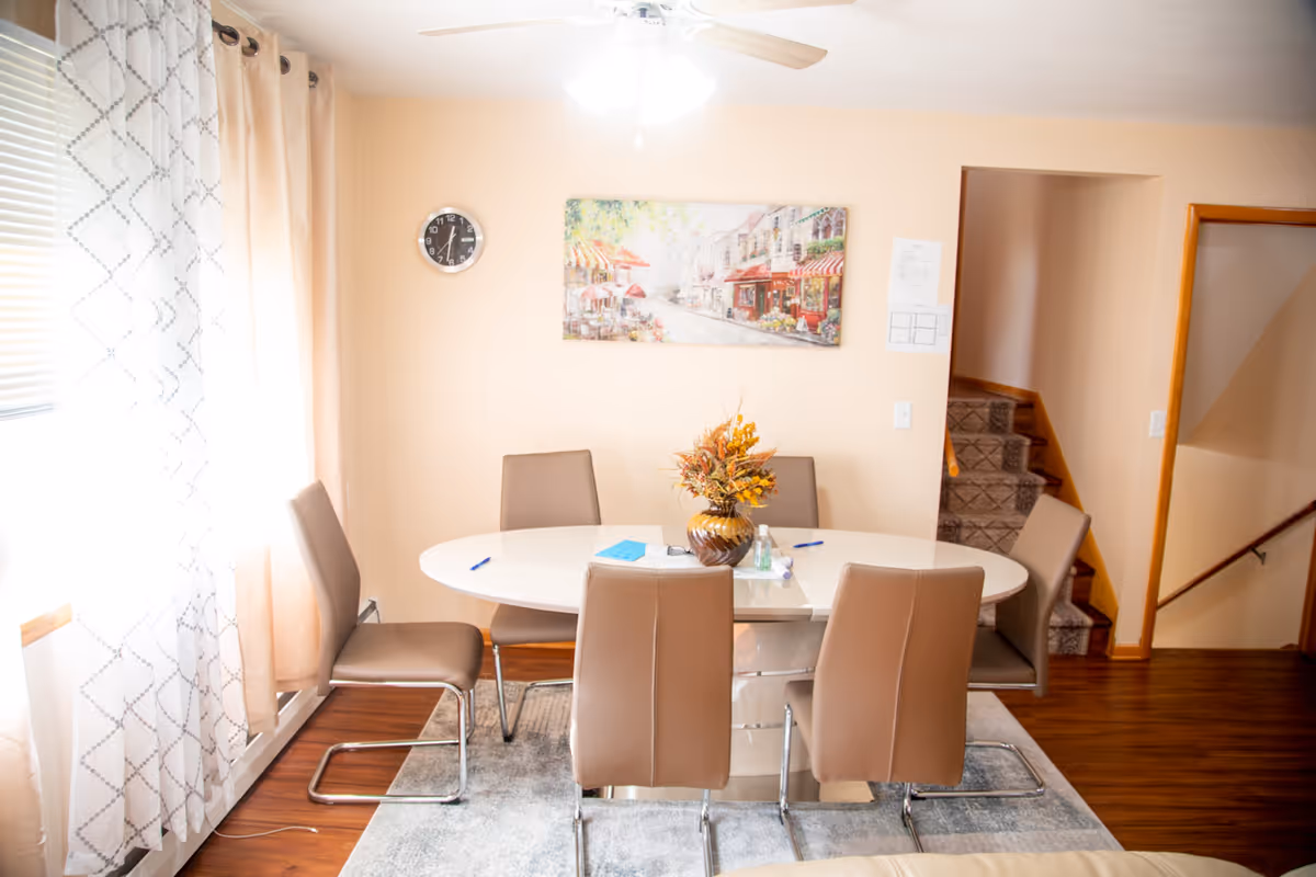 A round dining table with beige chairs, centerpiece, and sunlit windows in a residential dining room.