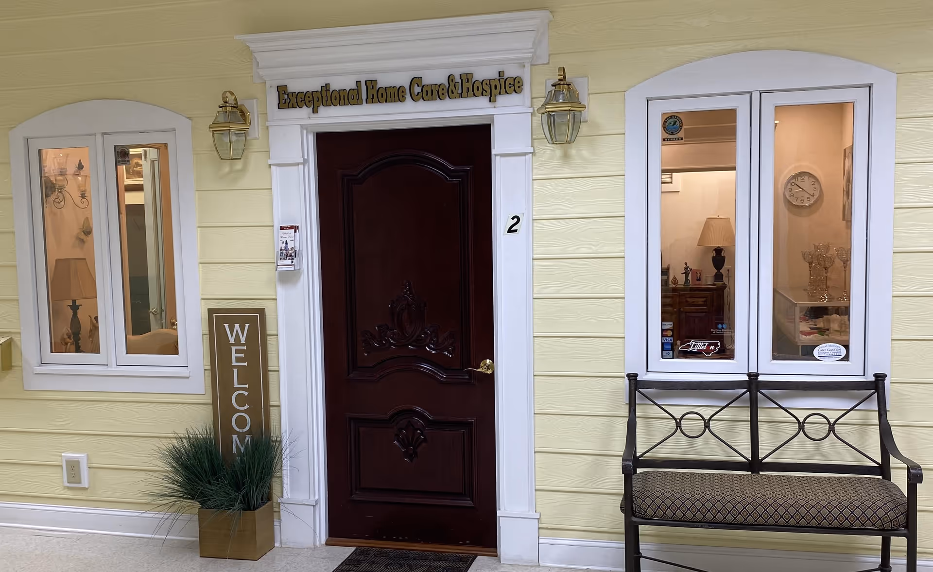 Cream-yellow exterior with a dark entry door, windows, bench, and a welcome sign.