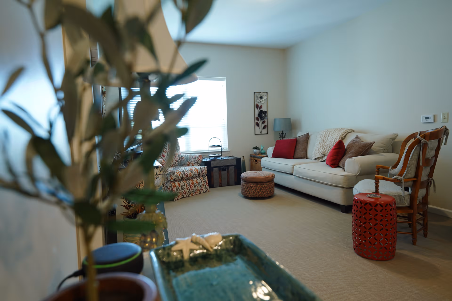 A cozy living room with a beige sofa adorned with red and brown pillows, a patterned armchair, a round ottoman, a wooden chair with a cushion, and a red decorative side table. The room has a window with blinds, a small lamp on a side table, and neutral-colored walls and carpet. A plant and a decorative blue dish are visible in the foreground.