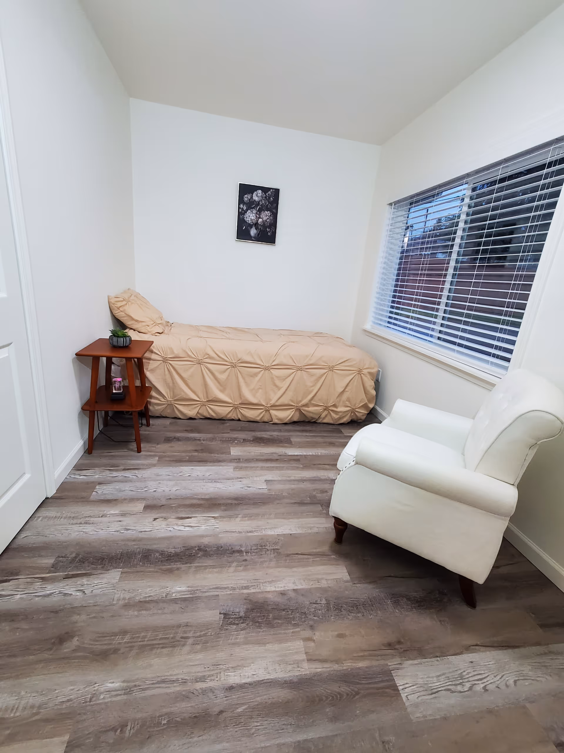 Small bedroom with beige bed, nightstand, and white armchair beside a window.