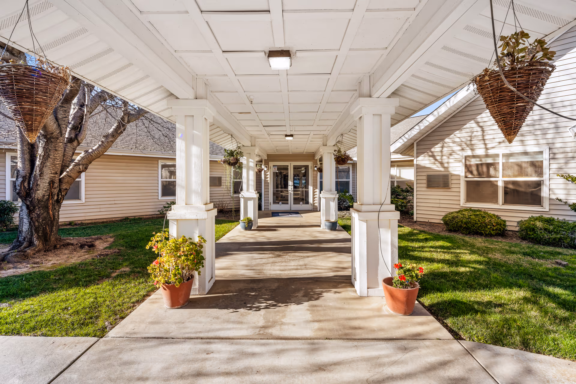 Covered walkway leading to a building entrance with white columns, hanging baskets, and planters.