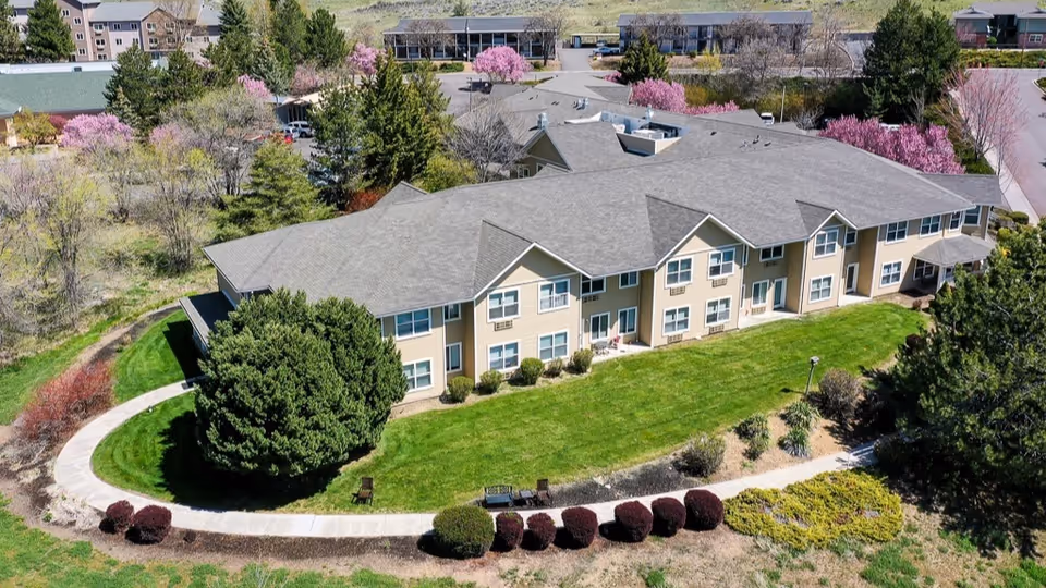 Aerial view of a senior living facility building surrounded by well-maintained green lawns, landscaped bushes, and trees with pink blossoms. A curved sidewalk runs along the front of the building, which has multiple windows and a gray roof. Additional buildings and parking areas are visible in the background.