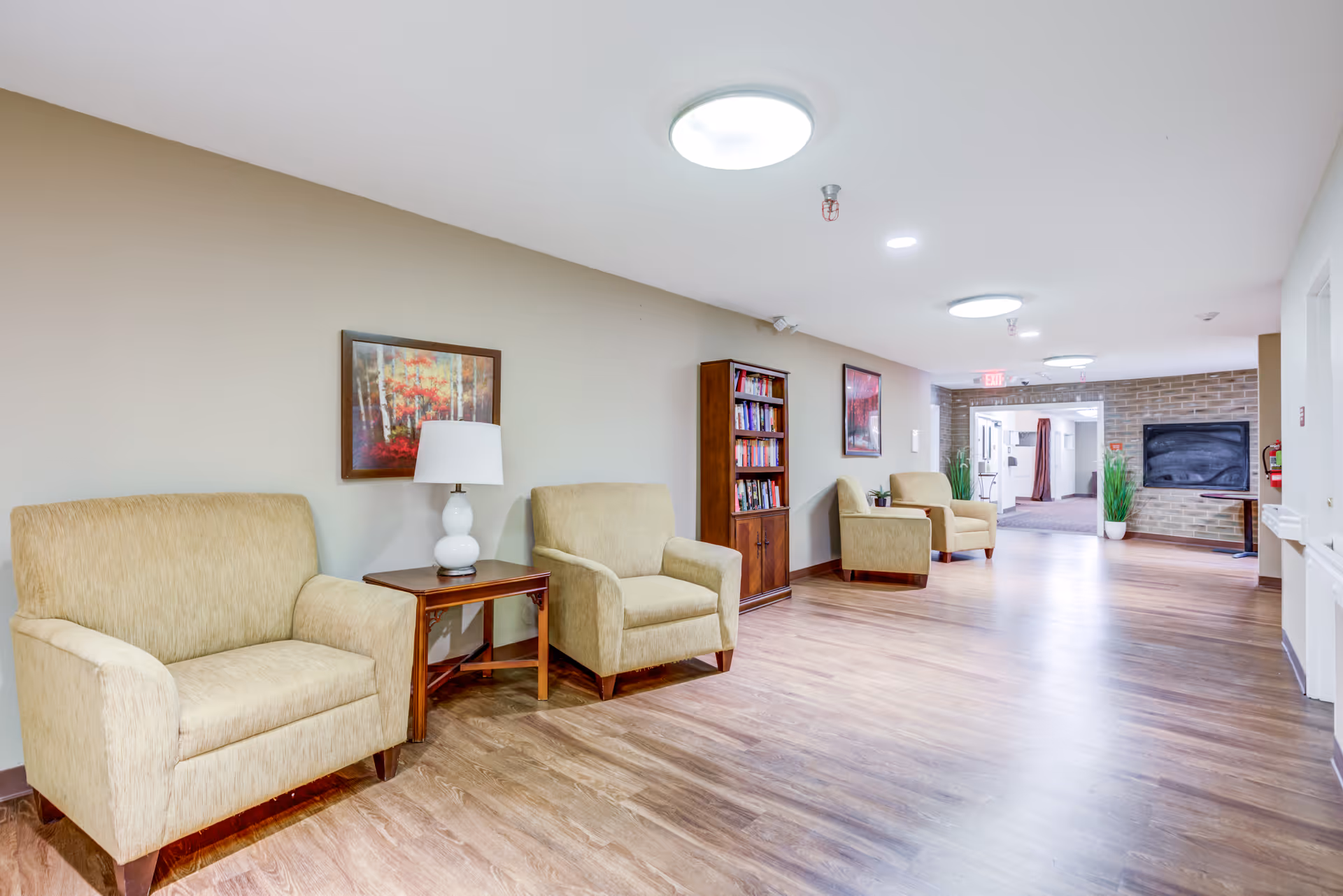 Bright senior living common area hallway with armchairs, side tables, a lamp, bookshelf and wood flooring.