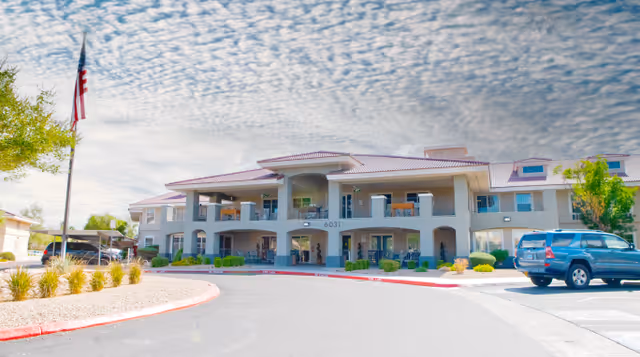 Exterior view of a two-story senior living facility building with balconies, a covered entrance, and a circular driveway. There is an American flag on a flagpole to the left and a blue SUV parked on the right side. The sky is partly cloudy with a textured cloud pattern.