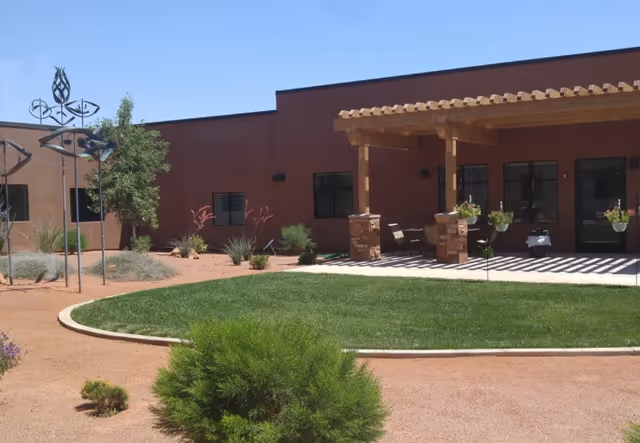 Outdoor courtyard area with a green lawn, desert landscaping, and a building with a shaded patio featuring stone pillars and hanging plants under a wooden pergola.