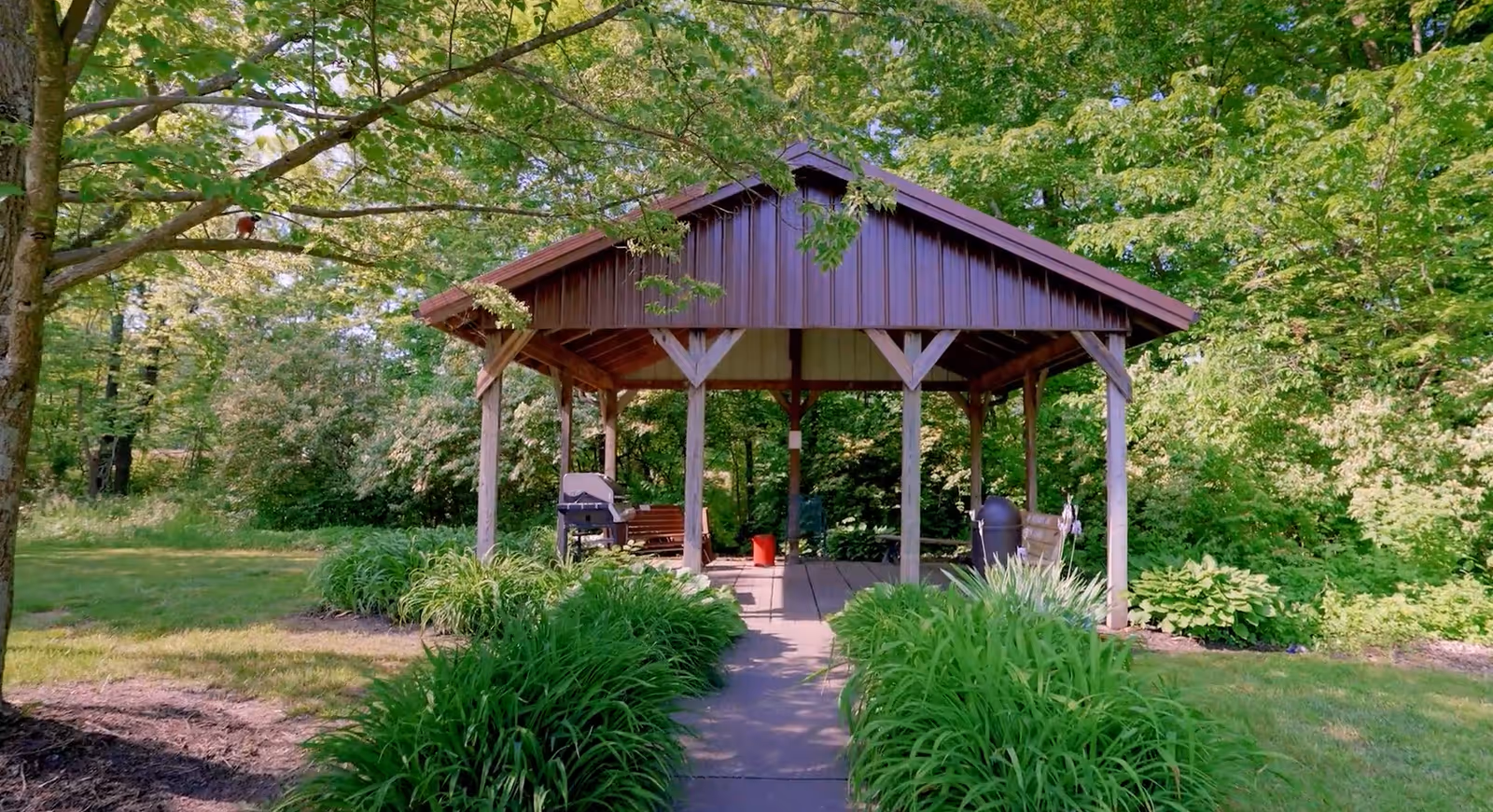 A wooden gazebo with a brown roof situated in a lush green garden area. The gazebo is surrounded by trees and bushes, with a paved pathway leading up to it. There are benches and a grill inside the gazebo.