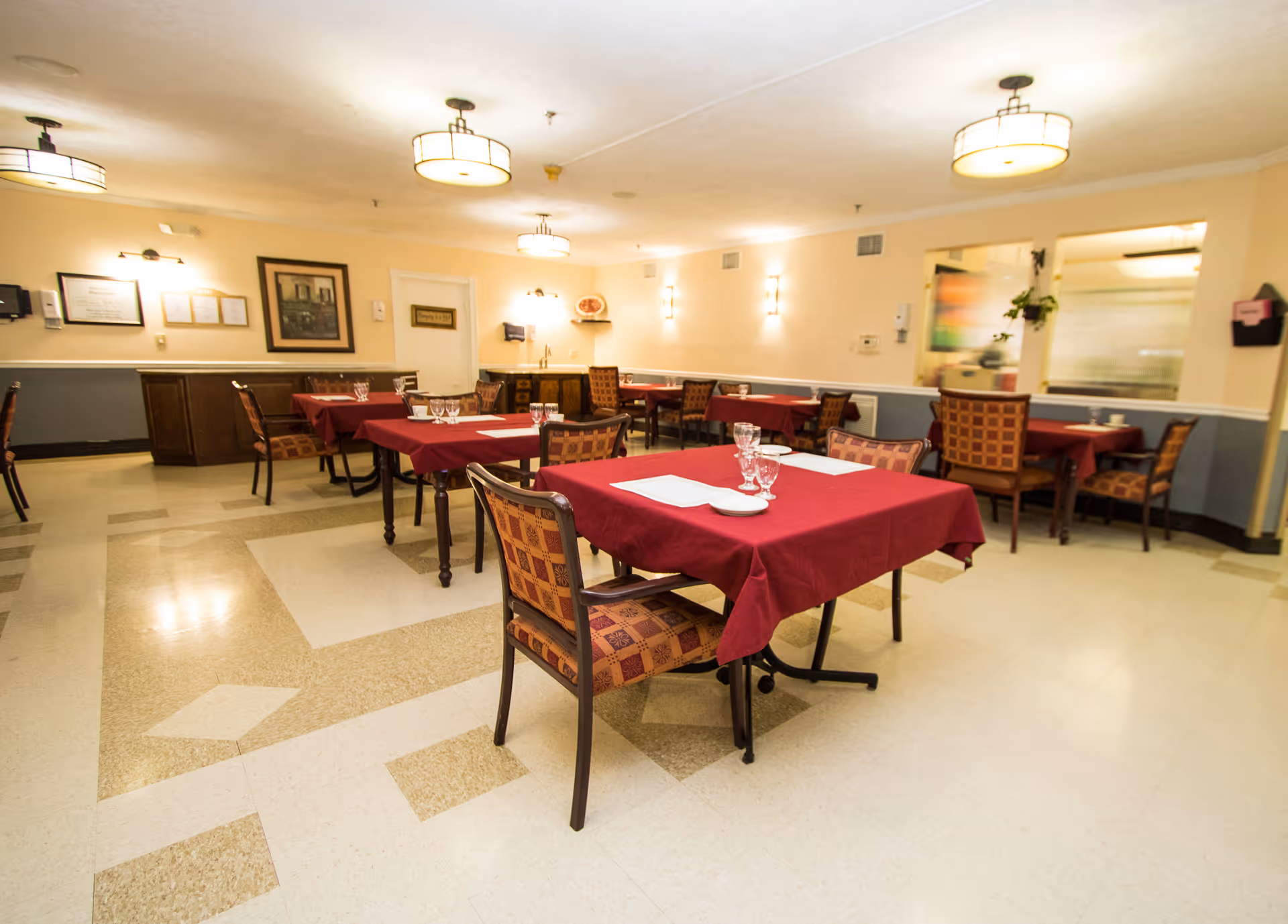 A dining room with several tables covered in red tablecloths, each set with glasses and napkins. The room has patterned chairs, beige walls with framed pictures, and ceiling lights providing warm illumination.