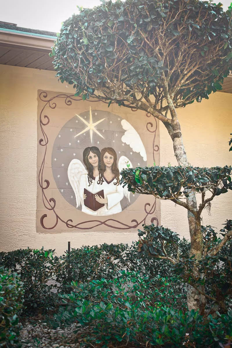 A mural painted on an exterior wall of a building depicting two angels with white wings holding a book, set against a circular background with stars and a large shining star above them. In front of the mural, there are neatly trimmed bushes and a tree with rounded foliage.