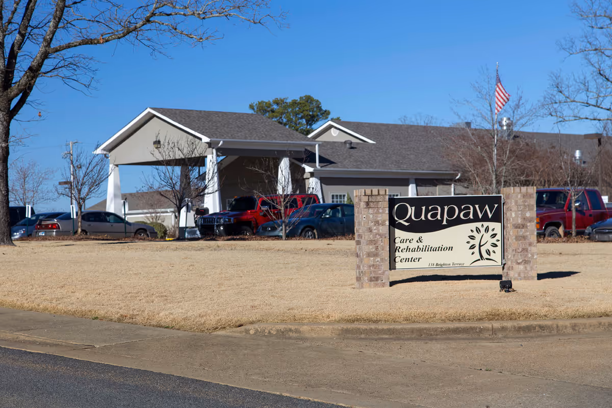 Exterior view of Quapaw Care & Rehabilitation Center building with a sign in front on a grassy area. Several parked cars and an American flag are visible under a clear blue sky.