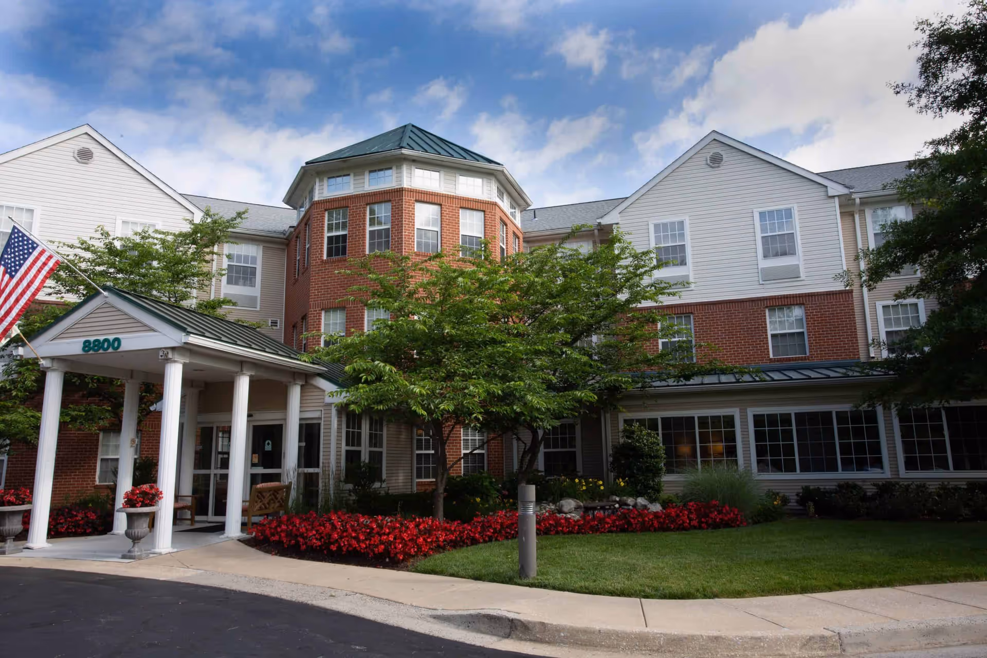 Exterior front entrance of a three-story senior living facility with columns, landscaped flowerbeds, and an American flag.