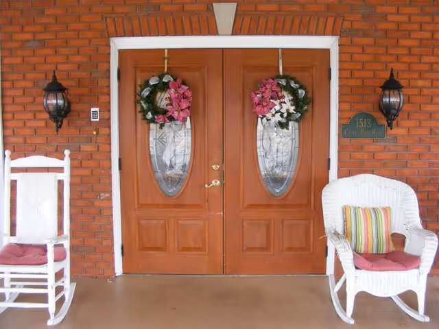 Double wooden doors on a brick entry with wreaths and white porch chairs.
