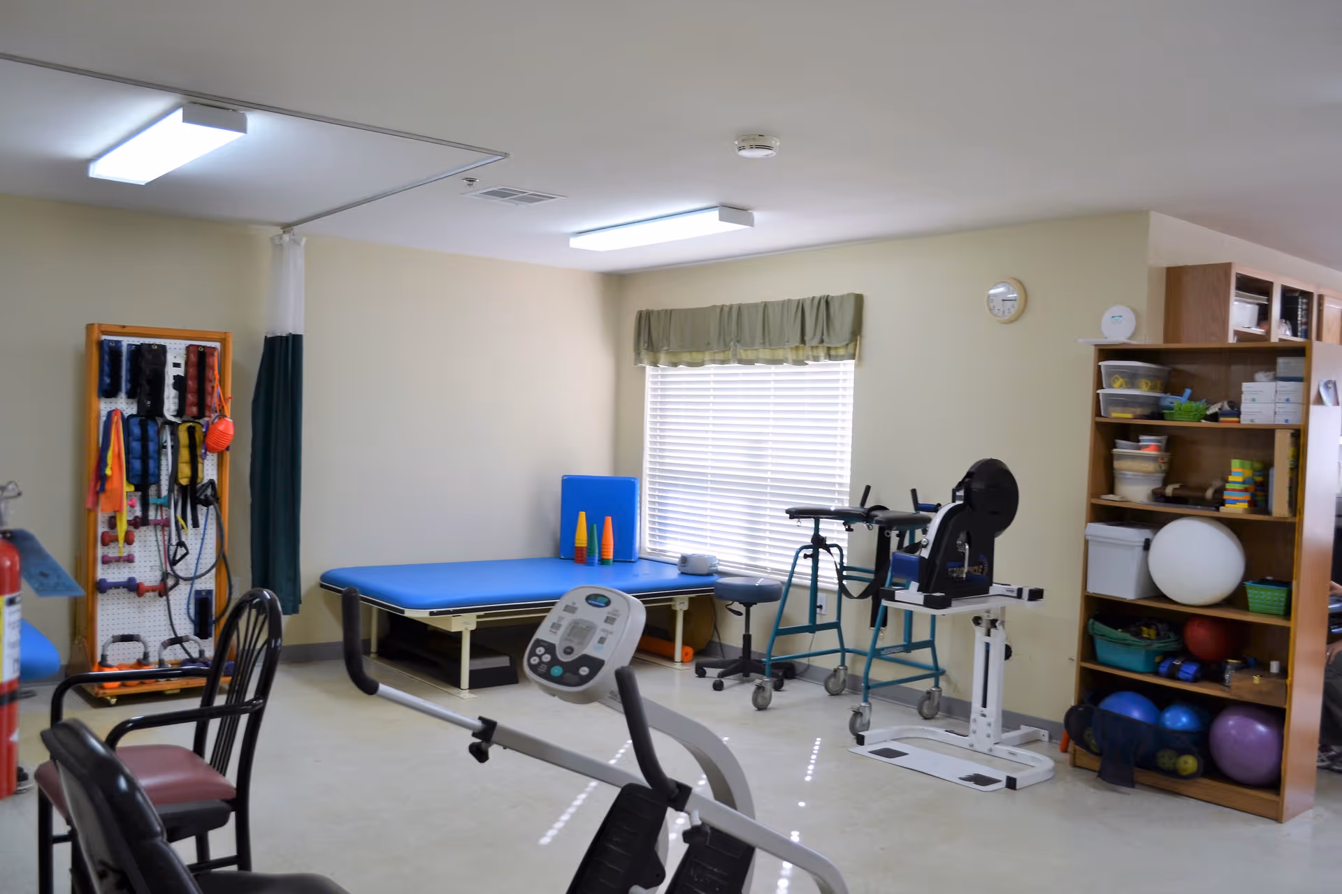 A therapy or exercise room with various rehabilitation equipment including a stationary exercise bike, a blue therapy table, a walker, and shelves stocked with exercise balls and other therapy tools. The room has beige walls, a window with blinds and a green valance, and fluorescent ceiling lights.