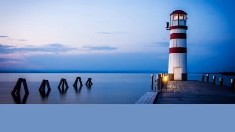 A striped lighthouse sits on a wooden pier over calm water at dusk.