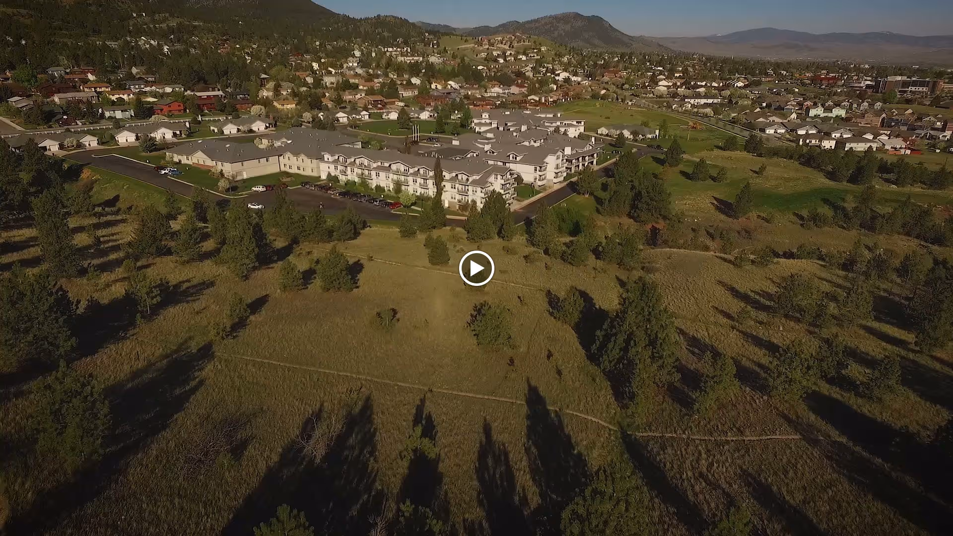 Aerial view of a senior living facility named Touchmark on Saddle Drive surrounded by green fields and trees, with a residential neighborhood and hills in the background under a clear sky.