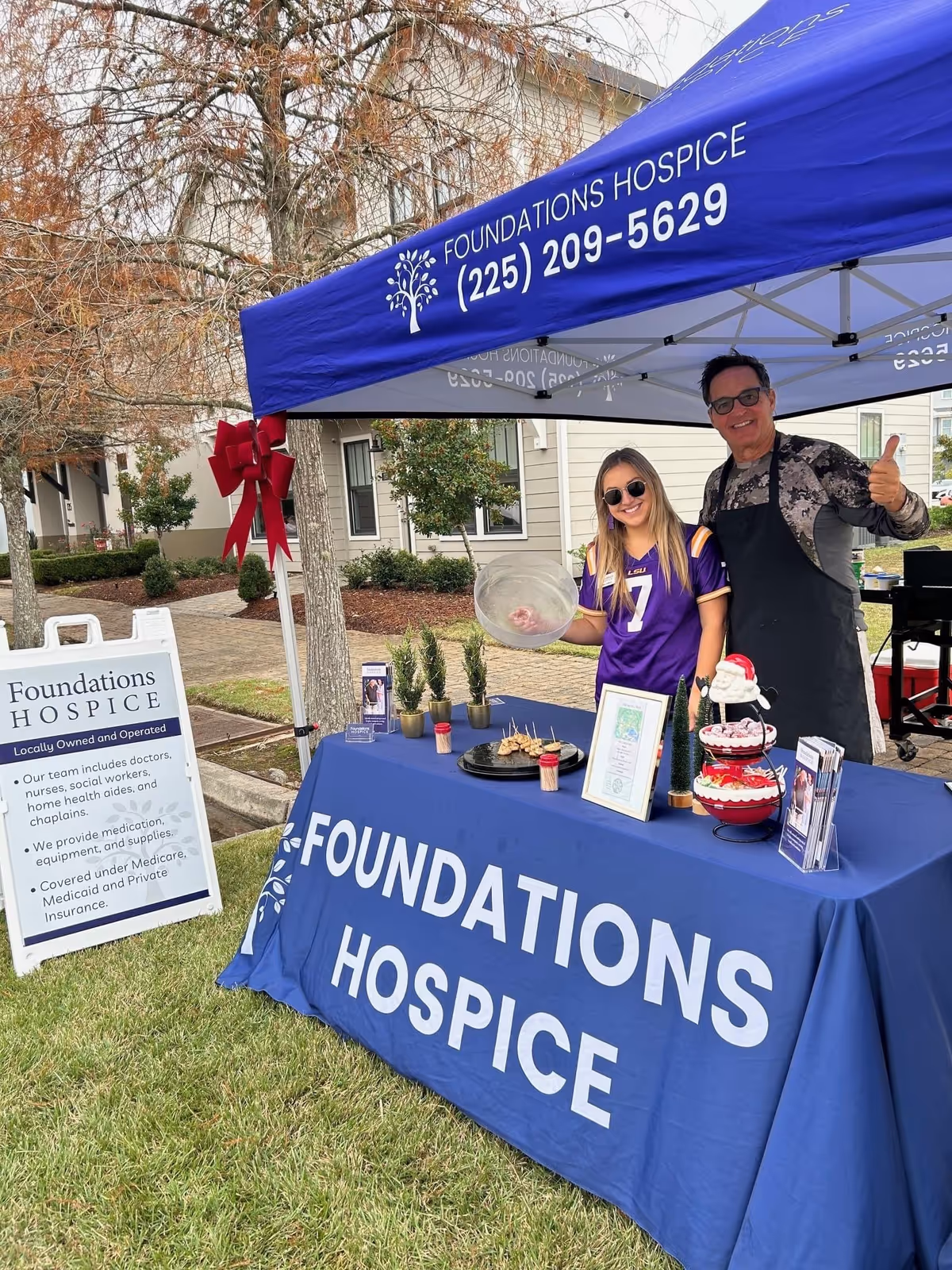 Two people stand at an outdoor Foundations Hospice tent with a long table and signage.